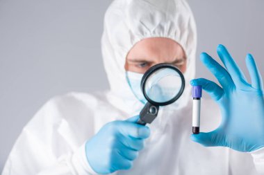 Solunum maskesi ve yüzünde korku olan mavi eldivenli koruyucu kıyafetli bir virüs uzmanı büyüteç ve kan dolu test tüpleri tutuyor. Salgın koronavirüs yüzünden sağlık sisteminin çökmesi.