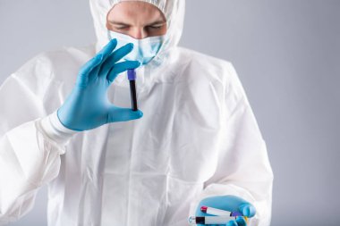 Tıbbi maskeli ve koruyucu giysili bir virüs uzmanı üzerinde koronavirüs testi için kan örneği olan bir test tüpü tutuyor. Salgın. Solunum sendromu, panik, deneyimler, araştırma
