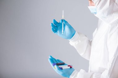 Solunum maskesi ve mavi eldivenli koruyucu kıyafetli virolog test tüplerinde şırınga ve kan testleri tutuyor.