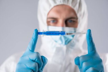 Solunum maskesi ve mavi eldivenli koruyucu kıyafetli virolog elinde şırınga tutuyor.