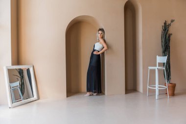 Duygusal, genç, sarışın, baştan çıkarıcı bir kızın portresi. Geniş, geniş bir odada, duvara yaslanmış, düşünceli.