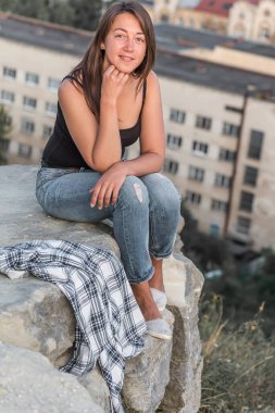 Gündoğumu genç, güzel, duygusal, kot pantolonlu, gömlekli ve siyah tişörtlü seksi kız şehrin arka planında bir kayanın üzerinde oturuyor.