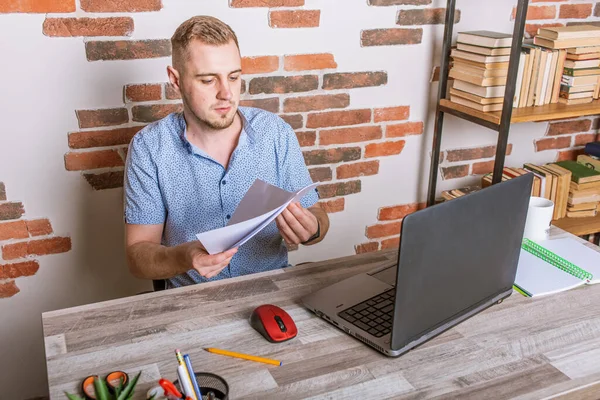 Young European man holding a piece of white paper in his hand, nervous, surprised and emotional, working at a computer with documents in a modern office, worried, worried about his work