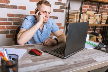 Genç erkek işadamı ofiste uzaktan çalışıyor, koltuklarda oturuyor bilgisayardaki belgelerle çalışıyor ve telefonla konuşuyor.