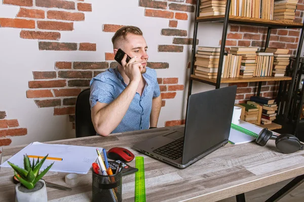 Genç erkek işadamı ofiste uzaktan çalışıyor, koltuklarda oturuyor bilgisayardaki belgelerle çalışıyor ve telefonla konuşuyor.