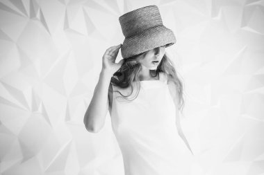 Aydınlık bir odada, açık renk bir elbise giymiş, hasır şapkalı genç bir kız. Seçici odak