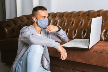 Gri maskeli genç bir adam koronavirüsün yayılmasını önlemek için karantina sırasında dizüstü bilgisayarıyla uzaktan çalışıyor. Adam COVID-19 salgını sırasında evden çalışıyormuş.. 