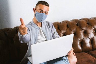 Gri maskeli genç bir adam koronavirüsün yayılmasını önlemek için karantina sırasında dizüstü bilgisayarıyla uzaktan çalışıyor. Adam COVID-19 salgını sırasında evden çalışıyormuş.