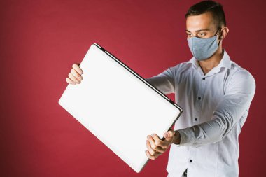 Gri yüzlü, koruyucu gri maskeli genç bir adam. Önünde beyaz dizüstü bilgisayarlı, kırmızı izole bir arka plan tutuyor. Coronavirus kontrol konsepti. Uzaktan karantina