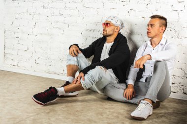 İki şık genç adam, biri bandana ve pembe gözlüklü, diğeri kot pantolonlu ve açık renk tişörtlü, yerde oturuyor ve fotokopi odasına bakıyor.