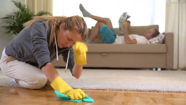 Unavená žena čistí parketové podlahy a její manžel ležel na pohovce s novinami