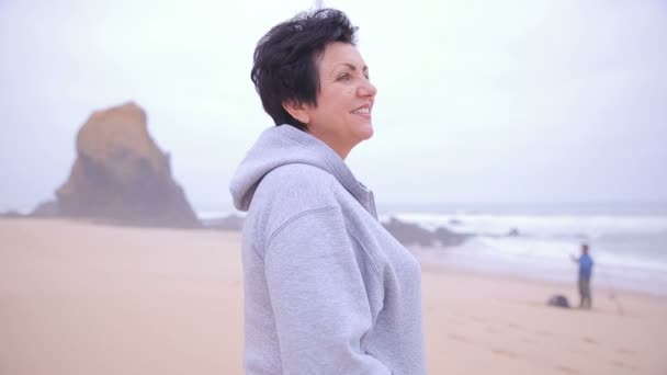 Attrayant actif mature femme debout sur une plage de l'océan et souriant à la caméra . — Video