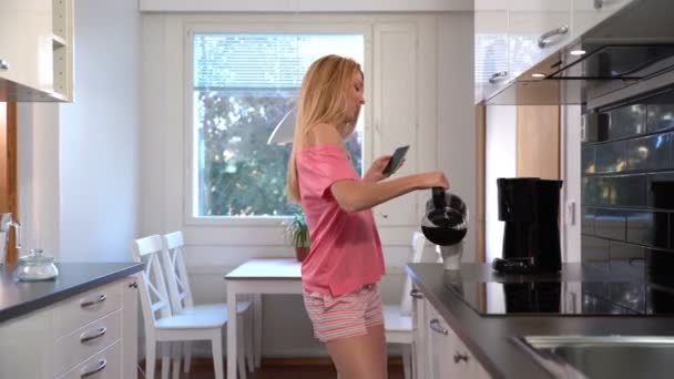 Een gelukkige vrouw in pyjama, dansen in de keuken, koffie, koptelefoon, smartphone. Dolly. — Stockvideo