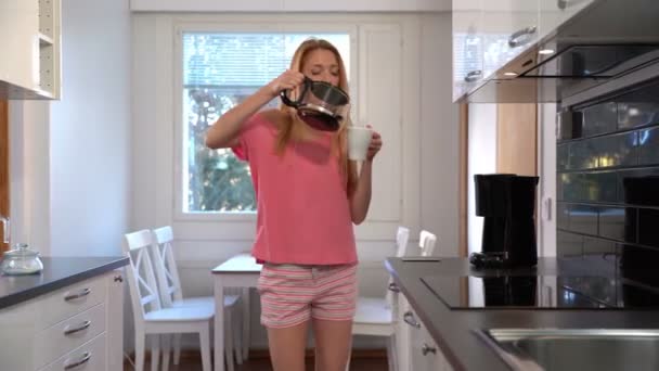 Een gelukkige vrouw in pyjama, dansen in de keuken, koffie, koptelefoon, smartphone. Dolly. — Stockvideo