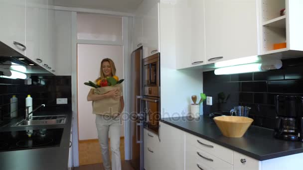 Gelukkig jonge vrouw brengt een zak met boodschappen naar de keuken. — Stockvideo