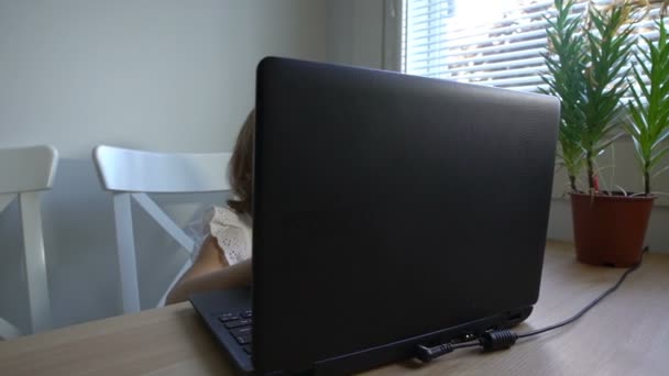 Little girl with interest uses the laptop sitting at the table. Dolly shot. — Stock Video