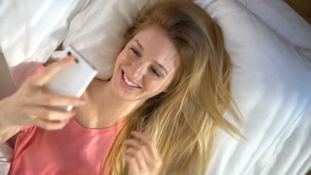 Sabahleyin yatakta yatarken, belgili tanımlık smartphone kullanarak ve gülümseyerek mutlu bir kadın — Stok video