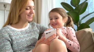 Genç anne ve kızı bir akıllı telefon çalmaya ve evde kanepede otururken gülümseyerek. Ağır çekim