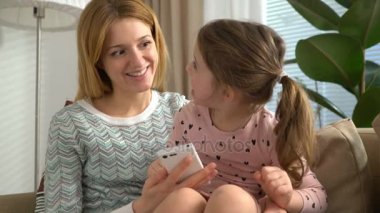 Genç anne ve kızı bir akıllı telefon çalmaya ve evde kanepede otururken gülümseyen