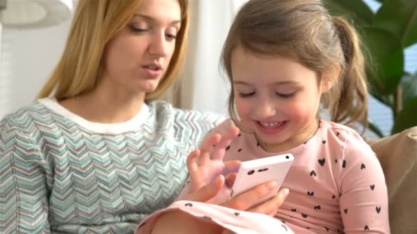 Νεαρή μαμά και κόρη είναι παίζοντας ένα smartphone και χαμογελώντας ενώ κάθεστε στον καναπέ στο σπίτι — Αρχείο Βίντεο
