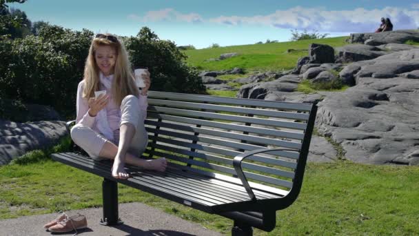 Una mujer joven utiliza un teléfono inteligente y bebe café en el banco en el parque. Movimiento lento . — Vídeo de stock