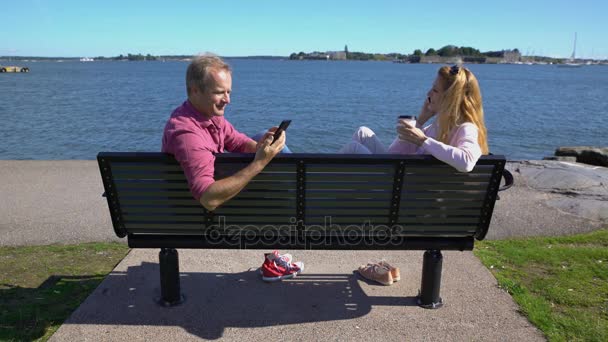 Una pareja sentada en un banco en la playa y usando sus teléfonos inteligentes — Vídeo de stock