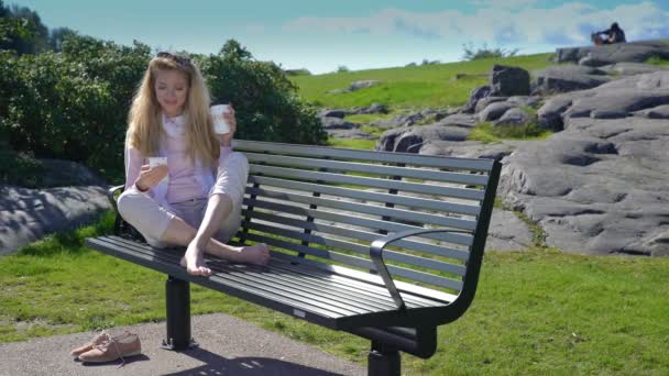 Uma jovem usa um smartphone e bebe café no banco no parque . — Vídeo de Stock
