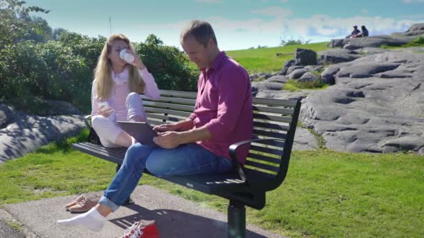 Jonge man met behulp van laptop, vrouw texting en drinken koffie in een stadspark. — Stockvideo