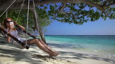 Genç bir kadın bir akıllı telefon kullanan ve tropik sahilde rahatlatıcı sahiptir.