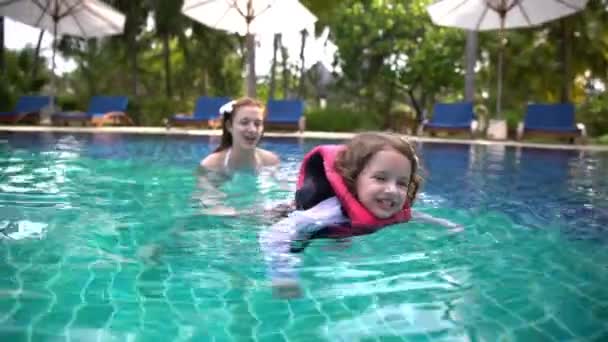 Маленькая девочка в плавательном жилете, плавающая в бассейне со своей матерью . — стоковое видео