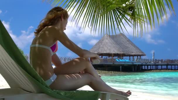 Jeune femme avec de belles jambes utilise de la crème solaire sur la plage tropicale. Mouvement lent — Video