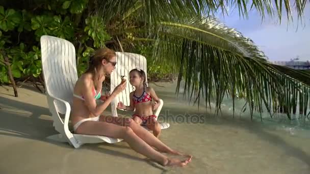 Eine junge Mutter und ihre kleine Tochter genießen Eis am tropischen Strand. — Stockvideo