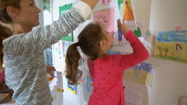 Niña con madre cuelga dibujos de niños en la pared en el vivero — Vídeos de Stock