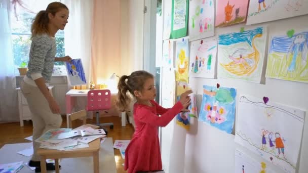 Niña con madre cuelga dibujos de niños en la pared en el vivero — Vídeos de Stock