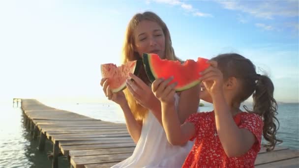 Glückliche Mutter und ihre Tochter sitzen auf einem Holzsteg und essen eine saftige Wassermelone — Stockvideo