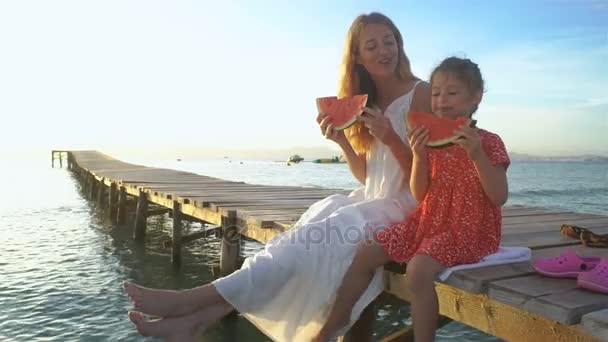 Feliz madre y su hija sentadas en un muelle de madera y comiendo una jugosa sandía — Vídeos de Stock