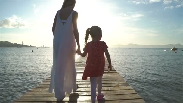 Mujer joven con su hijita caminando sobre un muelle de madera hacia el sol naciente — Vídeos de Stock