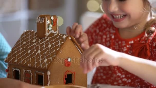 Junge Mutter und entzückende Tochter mit rotem Hut bauen zusammen Lebkuchenhaus. — Stockvideo