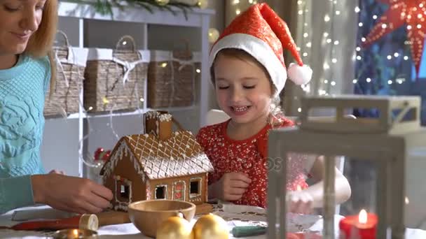 若い母親とジンジャーブレッドの家を一緒に造る赤い帽子でかわいい娘. — ストック動画