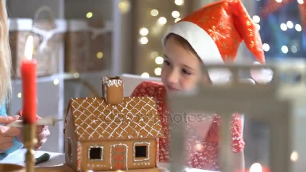 Young mother and adorable daughter in red hat building gingerbread house together. — Stock Video