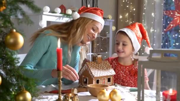 Young mother and adorable daughter in red hat building gingerbread house together. — Stock Video