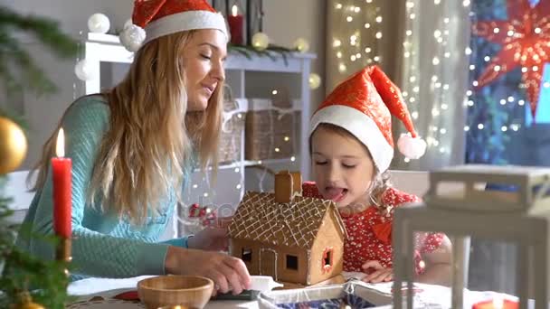 Young mom and cute child in red hat building gingerbread house together. Girl licking glaze off. — Stock Video