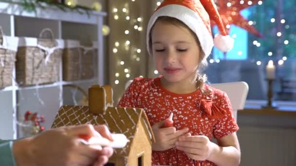 Junge Mutter und süßes Kind mit rotem Hut bauen zusammen Lebkuchenhaus. Mädchen leckt Glasur ab. — Stockvideo