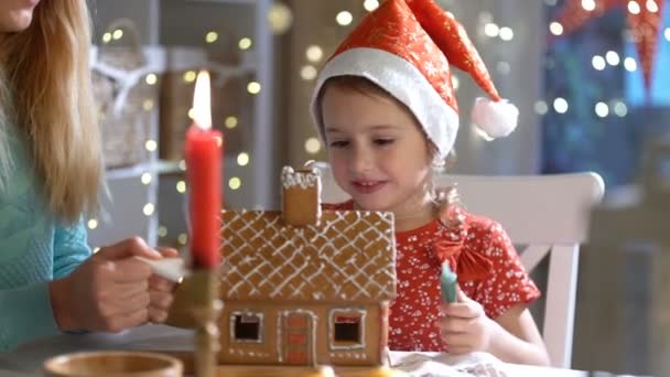 Young mother and adorable daughter in red hat building gingerbread house together. — Stock Video