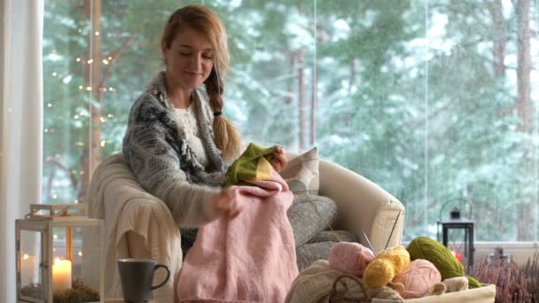 Молодая женщина вязала теплый шерстяной свитер в гостиной против снежного ландшафта снаружи — стоковое видео