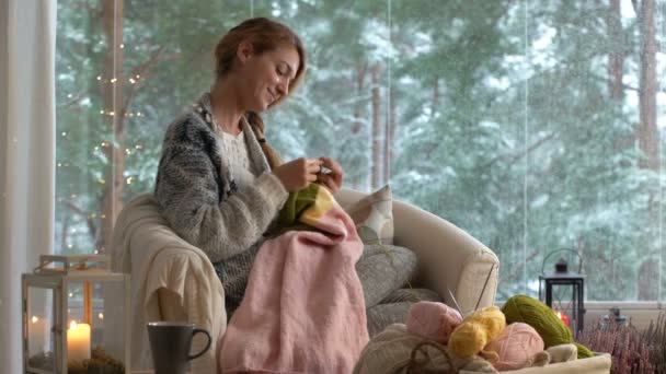 Νεαρή γυναίκα πλέξιμο ζεστό μαλλί πουλόβερ στο καθιστικό εναντίον του τοπίου χιονιού από έξω. — Αρχείο Βίντεο