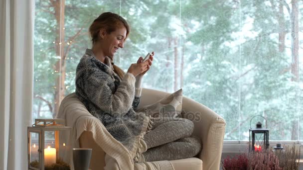 Eine Frau sitzt mit ihrem Smartphone in einem gemütlichen Wohnzimmer mit dem großen schneebedeckten Fenster — Stockvideo