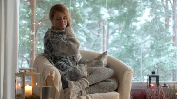 Затишний зимовий спосіб життя. Молода щаслива жінка п'є чашку кави в трикотажному светрі, сидить біля великого вікна з зимовим сніговим фоном — стокове відео