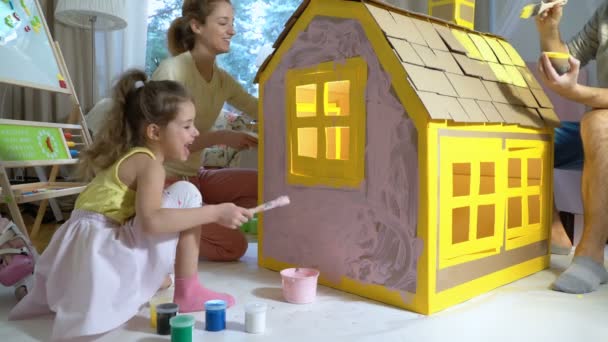 Família jovem com construção infantil e pintura de casa de papelão de brinquedo juntos . — Vídeo de Stock