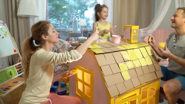 Famiglia giovane con la costruzione del bambino e la pittura giocattolo casa di cartone insieme . — Video Stock